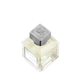Square glass perfume bottle with pale yellow liquid, a white label, and a distressed grey square cap with an embossed 'MK' lo