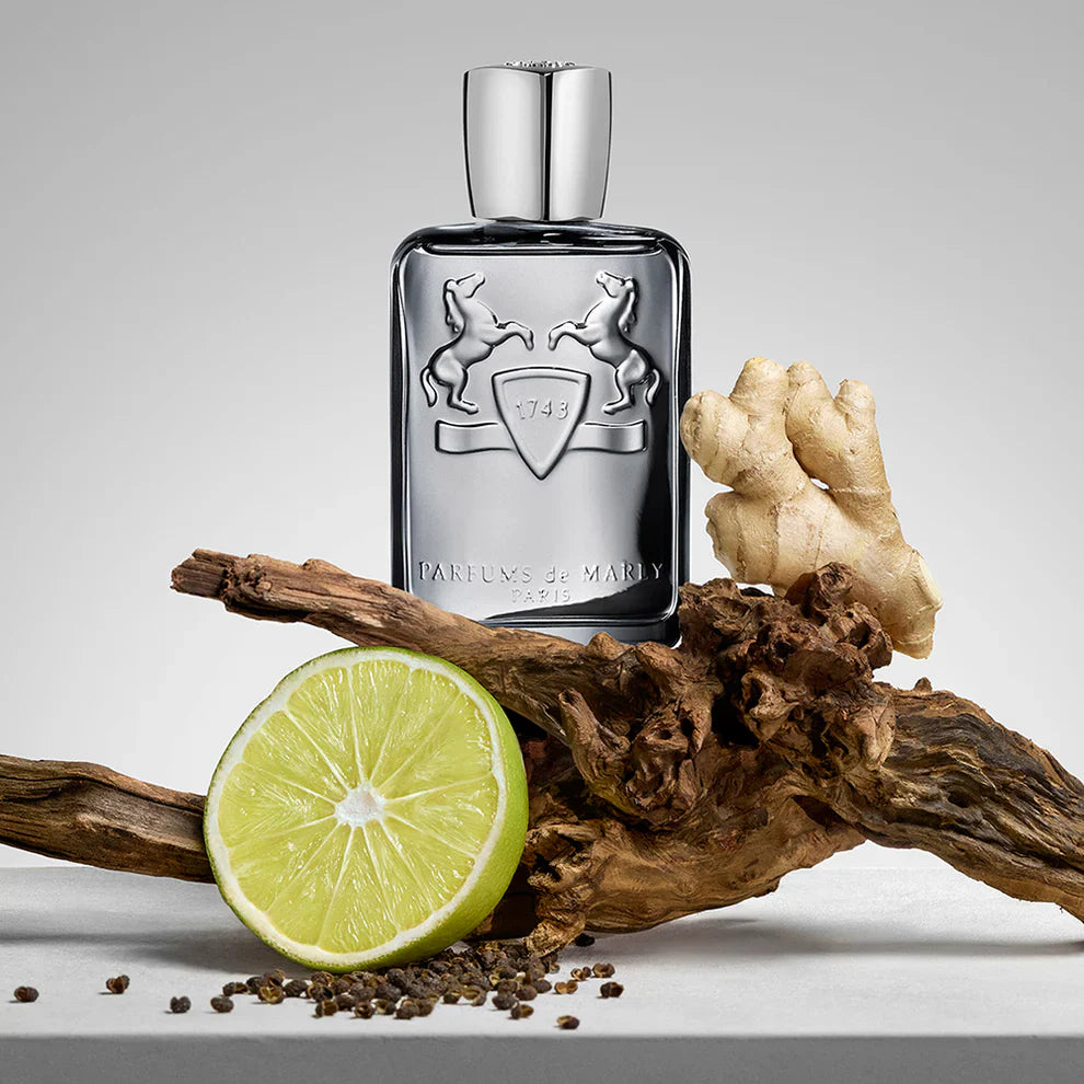 A metallic silver Parfums de Marly fragrance bottle artfully arranged with raw ingredients: a sliced green lime, a piece of ginger root, peppercorns, and textured driftwood.