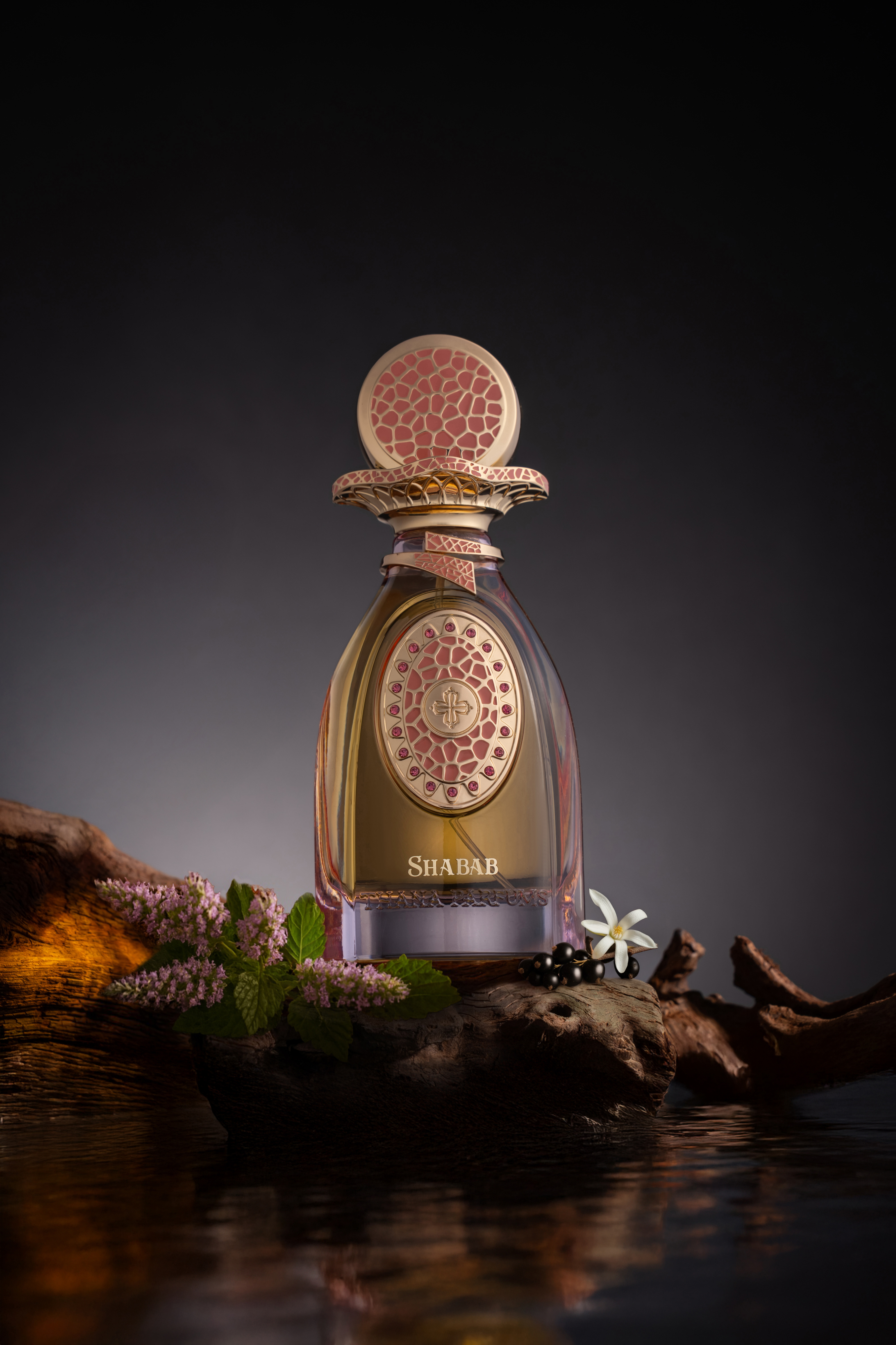 Clear glass perfume bottle with gold-toned liquid. Gold-toned cap, collar, and front emblem with pink mosaic, pink gems, and
