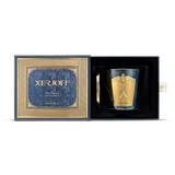 An iridescent, dark-toned glass candle holder with vibrant blue and green shifts, adorned with a large gold Xerjoff crest, shown next to a polished gold lid.
